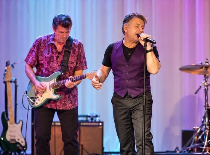 J.C. Grigg singing on stage in a purple vest while a guitarist plays beside him